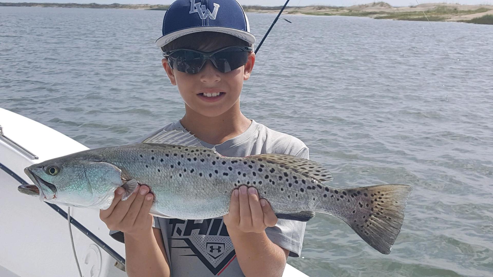 Boy on a boat smiles while holding a speckled trout he caught; ocean background.