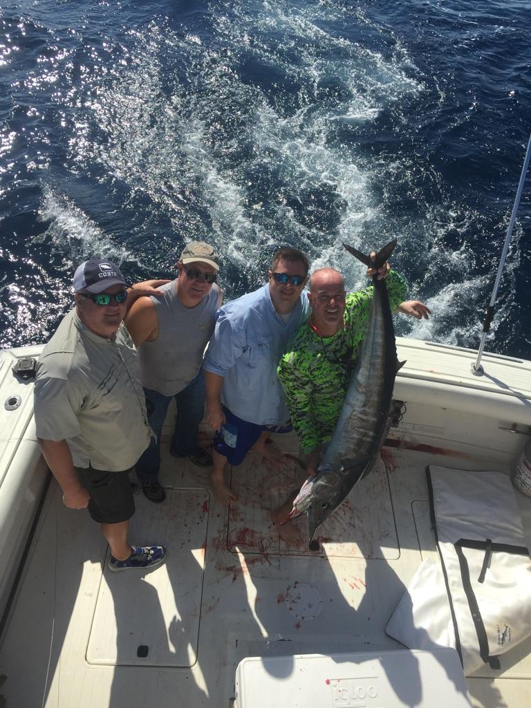 Four men on a boat, posing with a large fish they caught. Blue water in background.