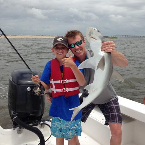 Father and son on boat, holding shark. Son gives thumbs up, smiling. Gray water and sky.