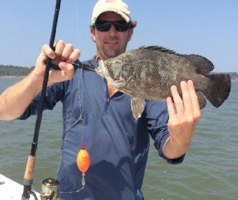 Man on a boat holding a dark fish he caught; fishing rod and bobber in view; sunny day.