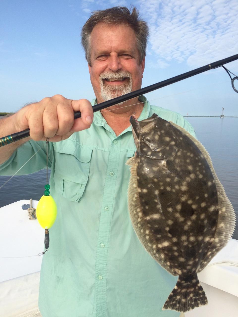 Man on a boat holding a flounder he caught, with fishing rod and bobber in a calm water setting.