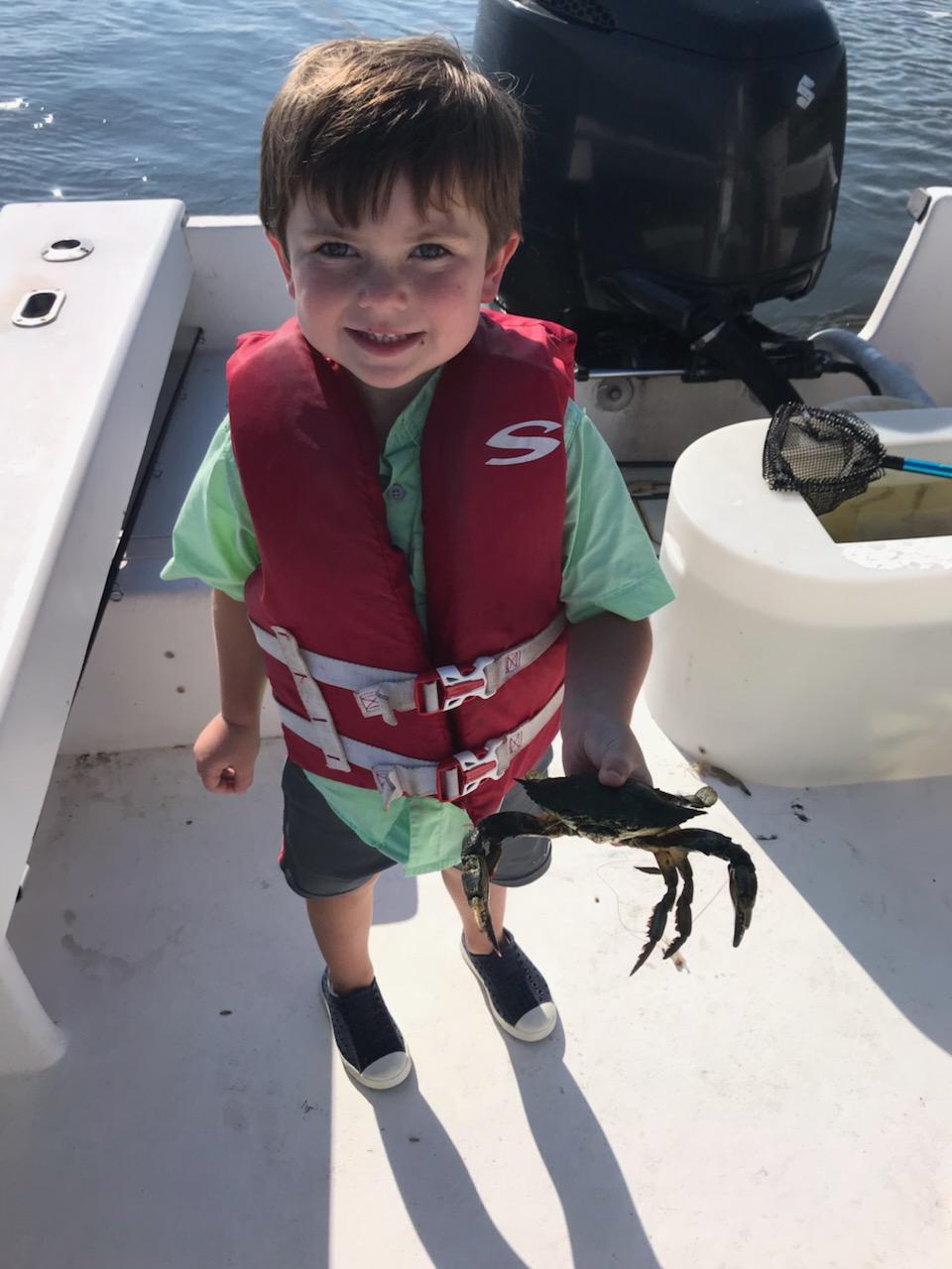 Boy in a life vest holding a crab on a boat, smiling, in sunlight.