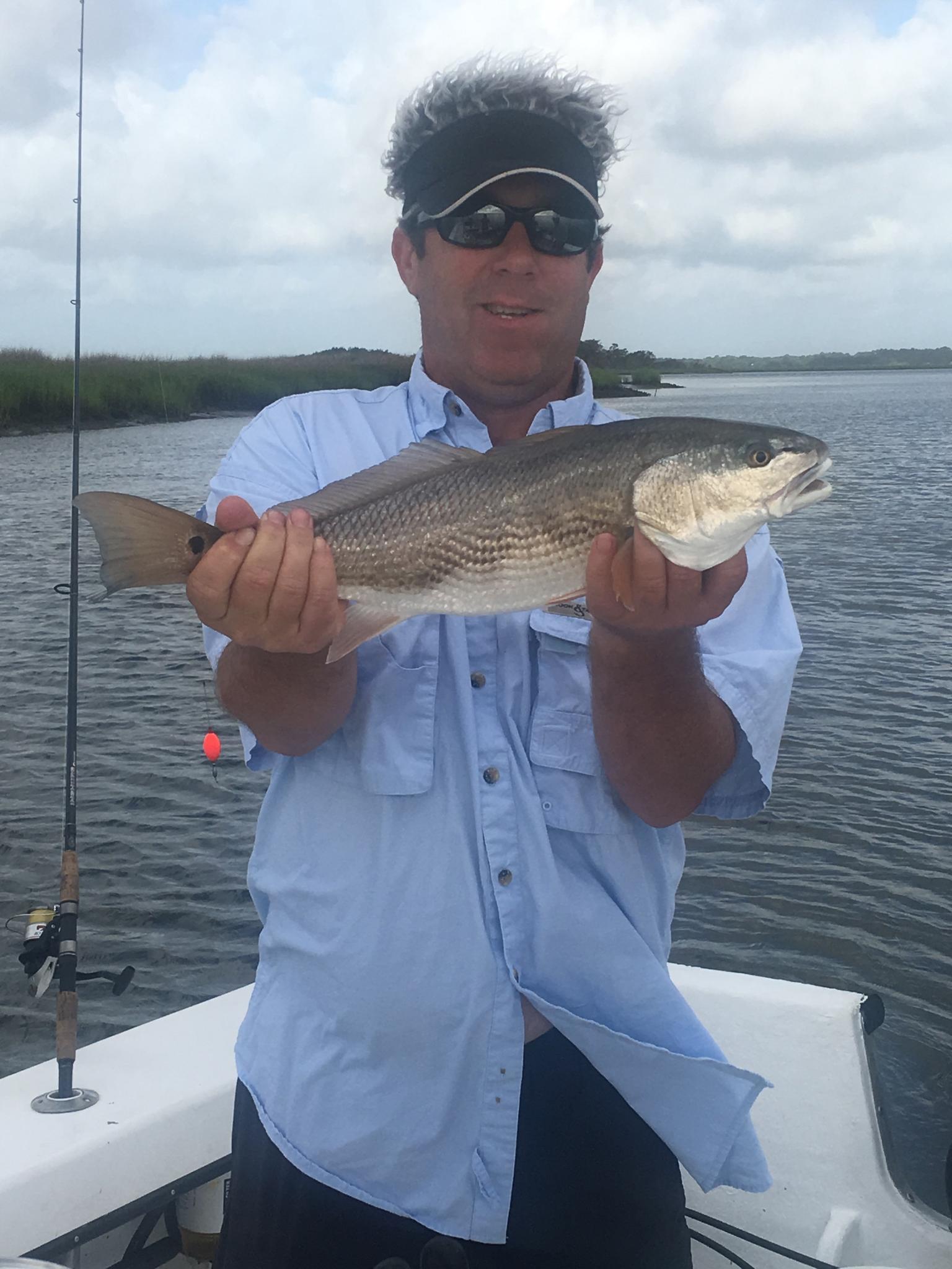 Man holding a fish on a boat. The fish is gray and silver. Setting is near water, cloudy sky.