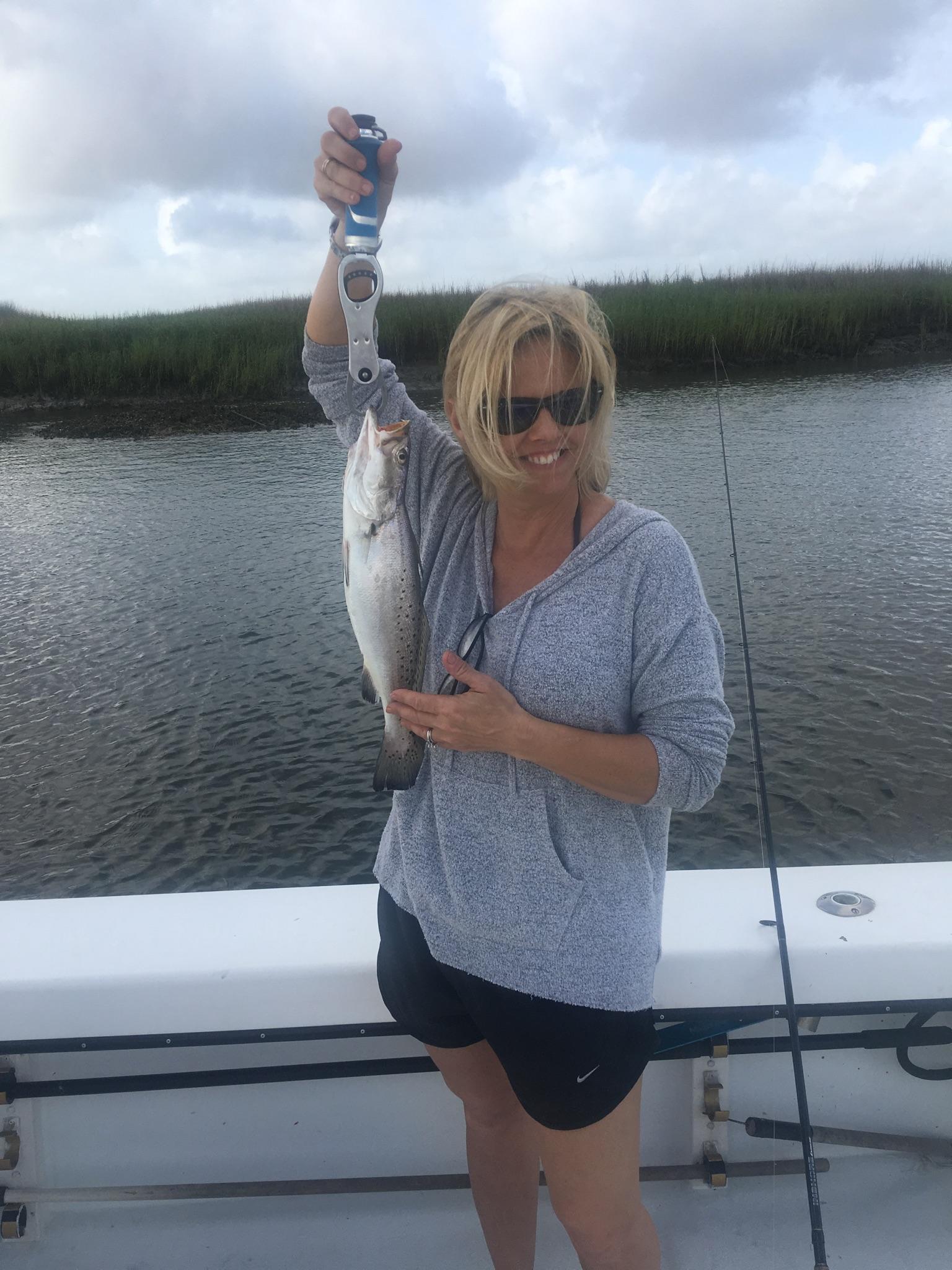 Woman on a boat holding a speckled trout, smiling, near a marsh.