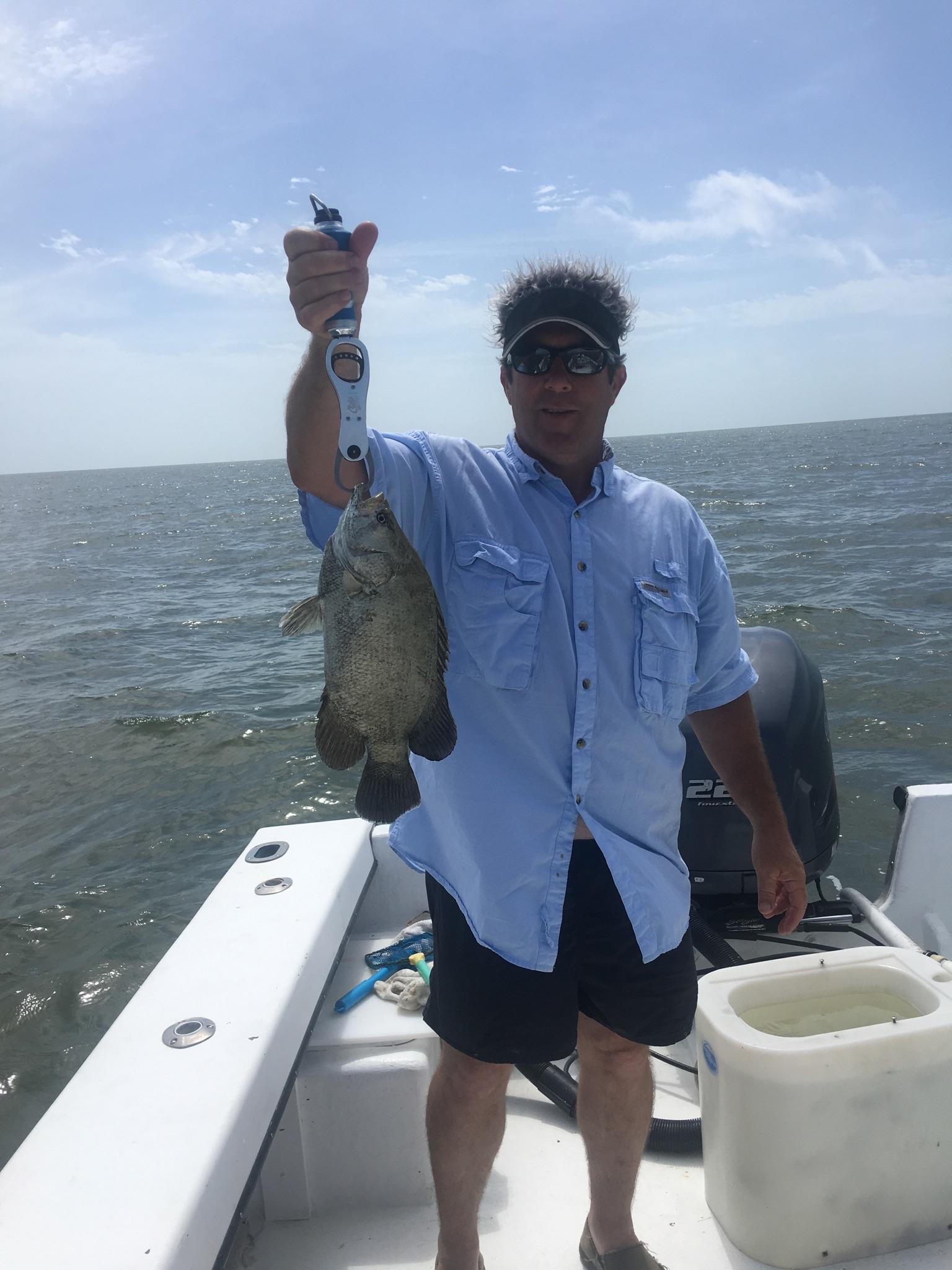 Man on boat holds up a spotted fish on a scale, blue sky and water.