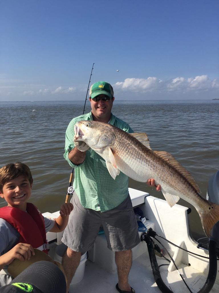 Man holding large redfish on a boat, smiling with a boy in a life vest. Ocean backdrop, sunny day.
