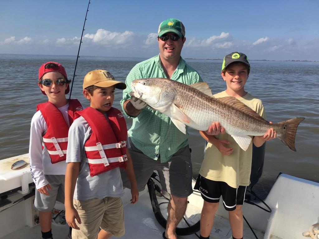Man and three boys on boat hold up large redfish. Blue water and sky.