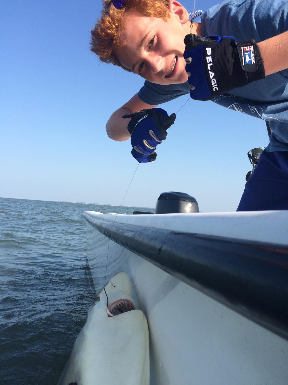 Boy smiles while reeling in a shark next to a boat on the water.