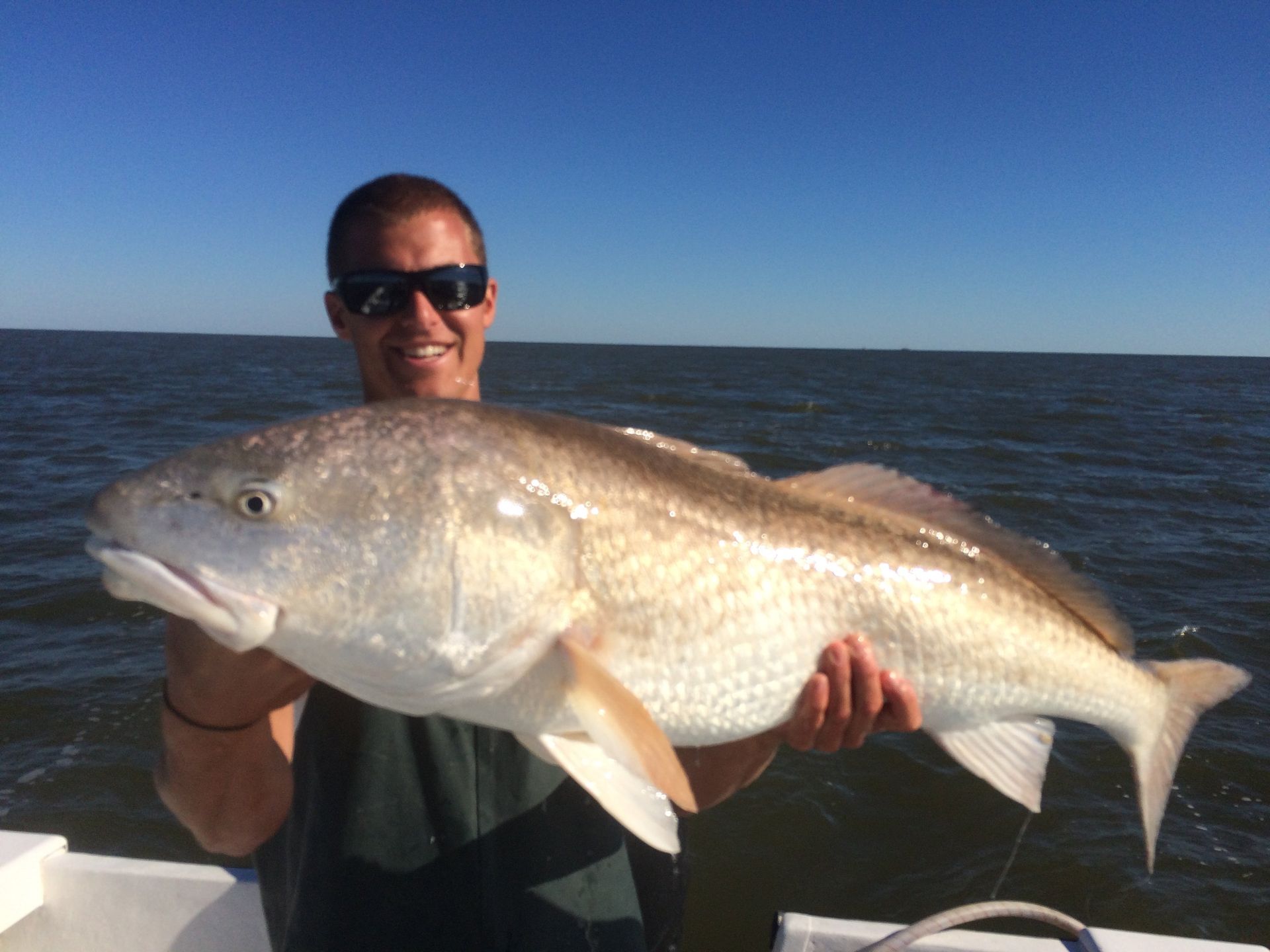 Man holding a large, reddish-bronze fish on a boat, smiling, with a blue sky and water.