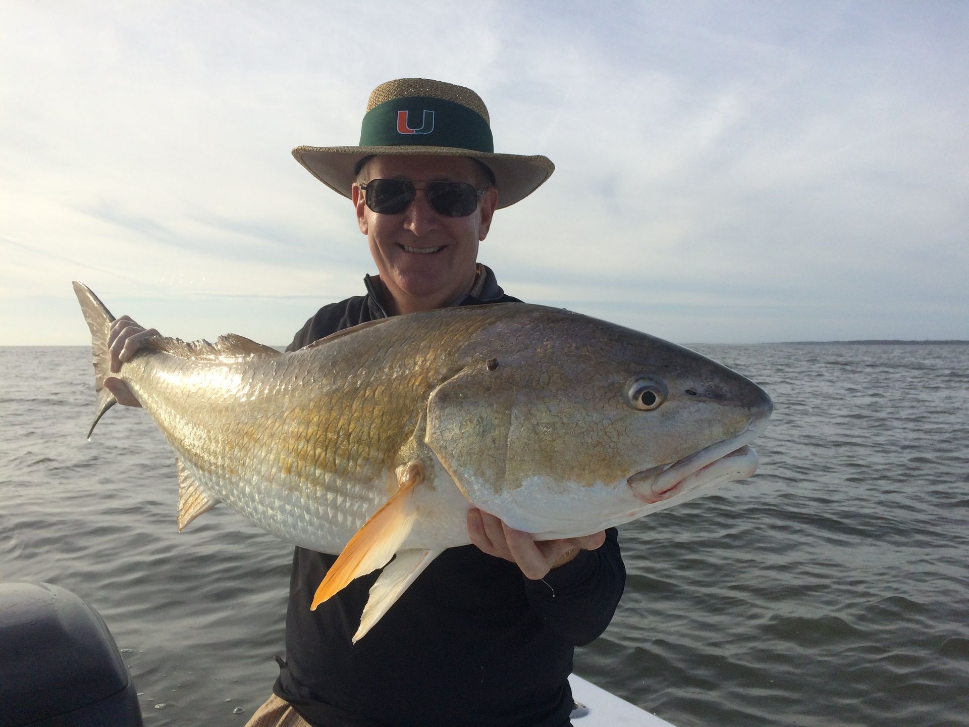 Man in sunglasses and hat holding a large redfish on a boat, near water.