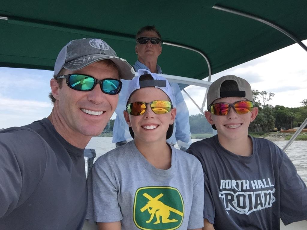 Four people smiling on a boat: two children, a man in a cap, and another man in the background.