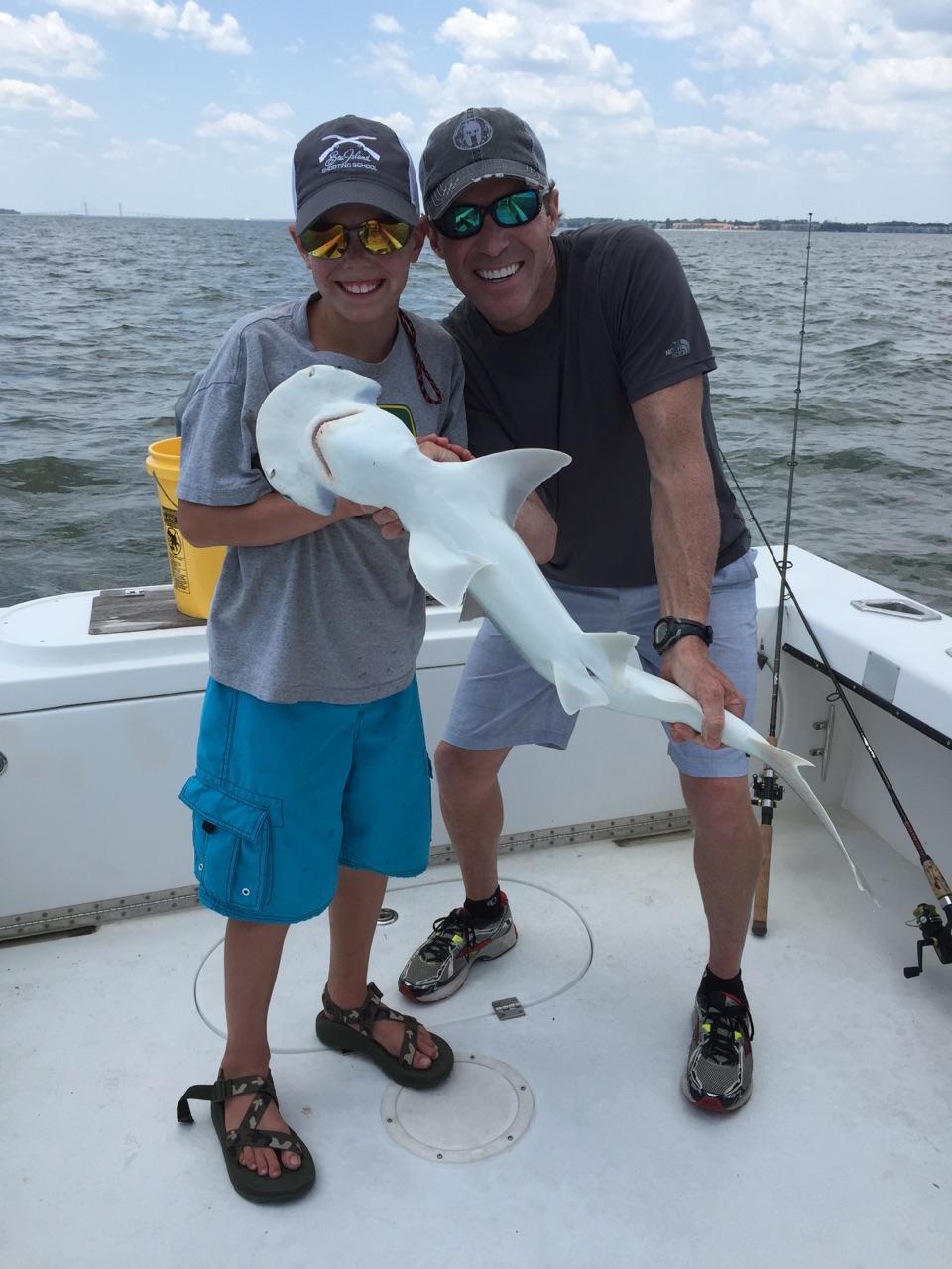 Father and child on a boat, holding a small white shark, smiling. Overcast day.