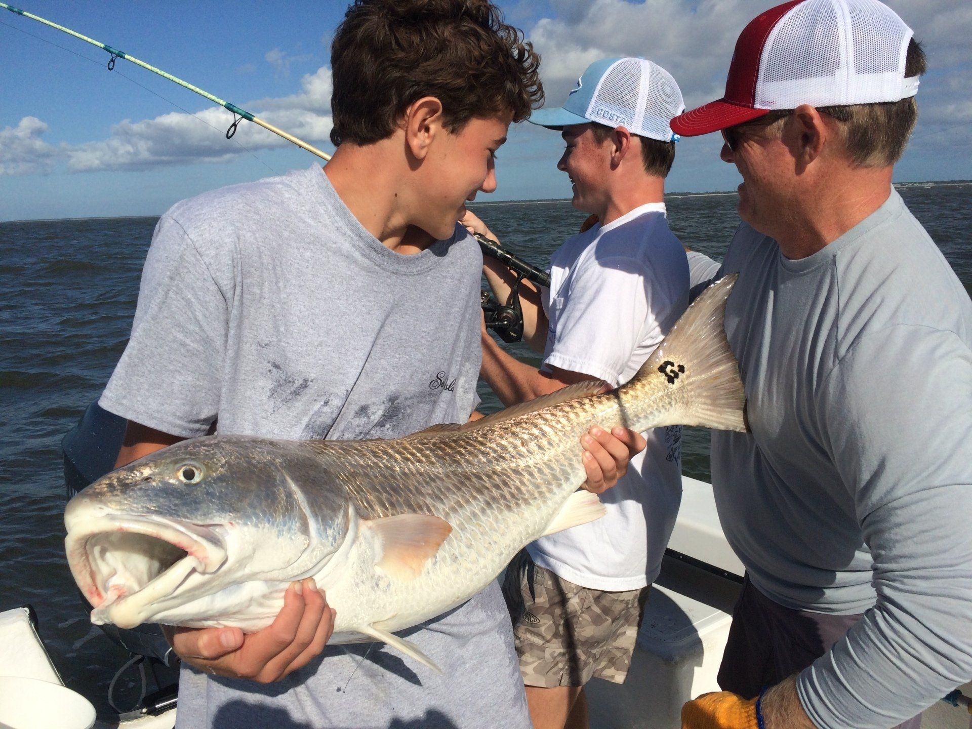 Three people on a boat holding a large redfish with its mouth open.  Smiling,  gray sky, ocean water.