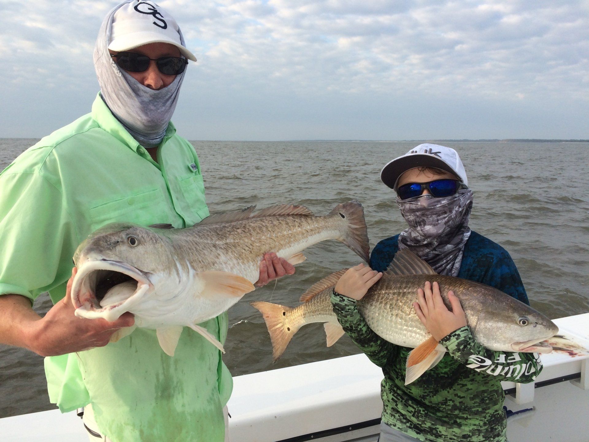 Two people on a boat holding large redfish. Both wear hats and face coverings; cloudy sky, water.
