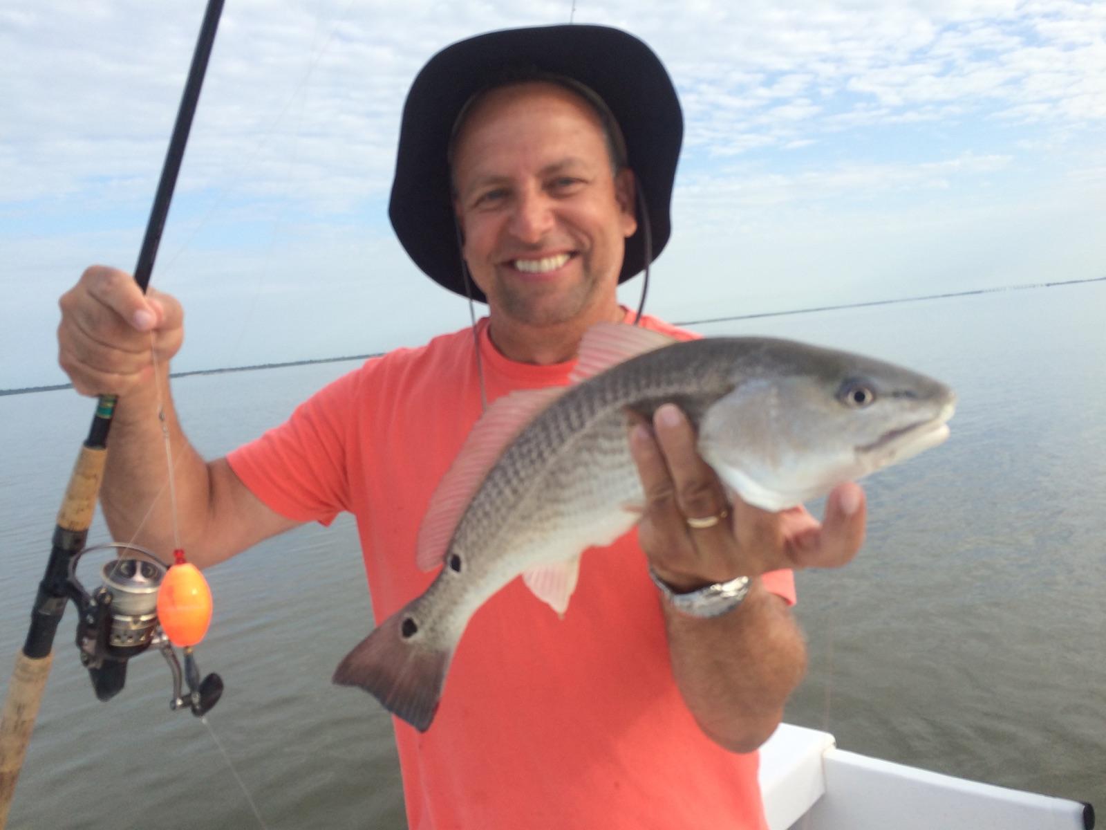 Man on boat holds up a redfish, smiles; fishing rod in hand, open water.