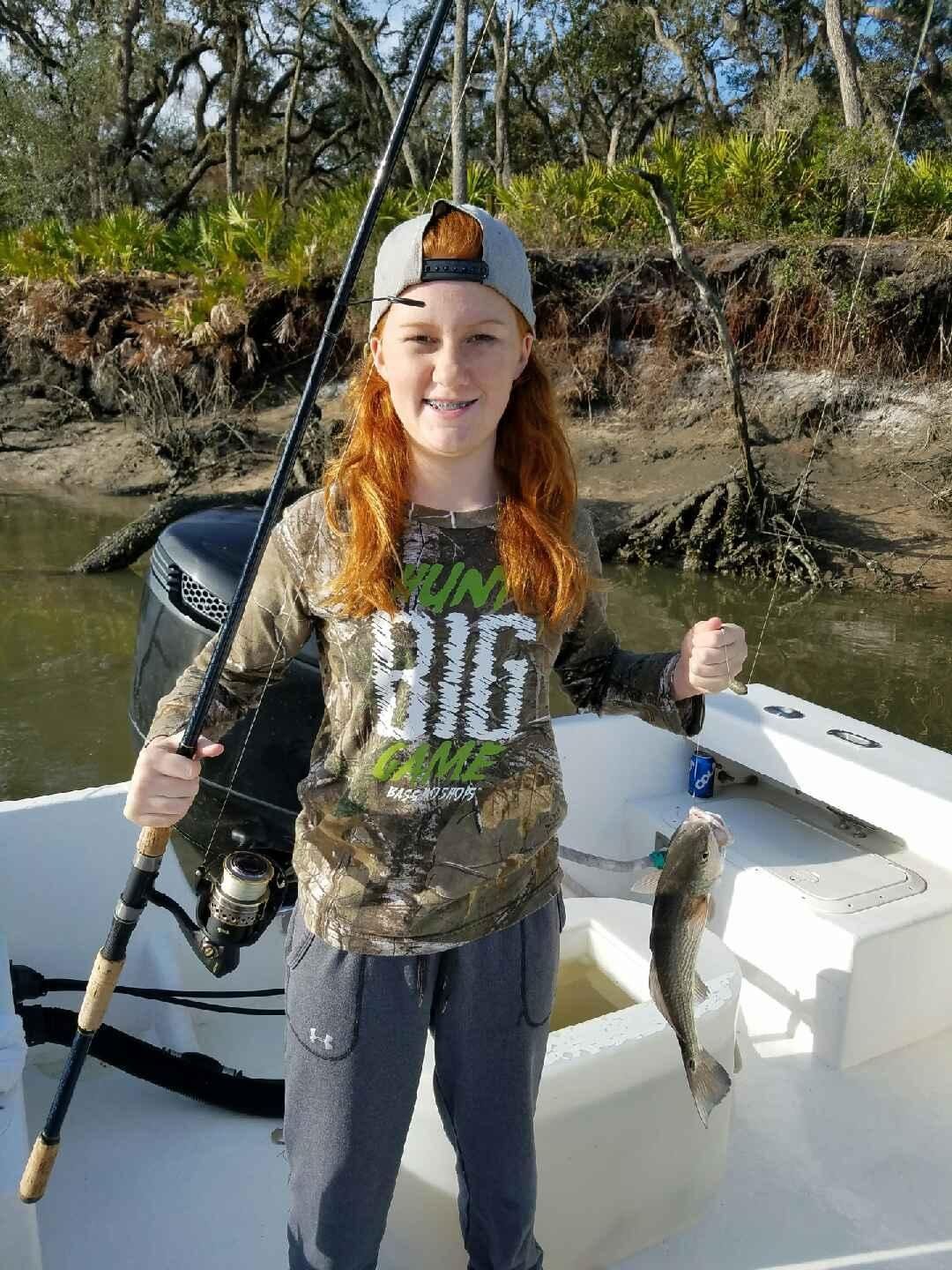Girl with red hair holds up a fish on a fishing rod on a boat, smiling, trees in background.