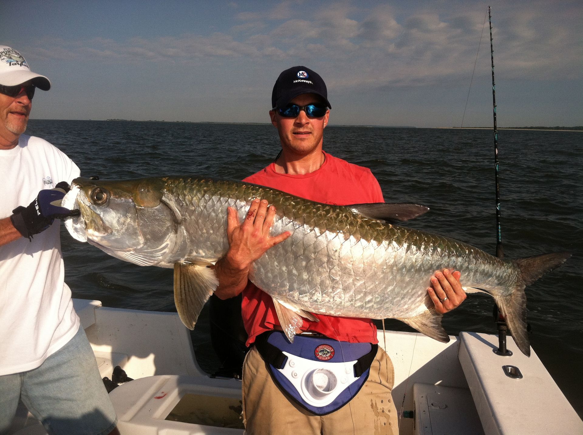 Man holding large silver tarpon fish on a boat. Another man helps, both near water.