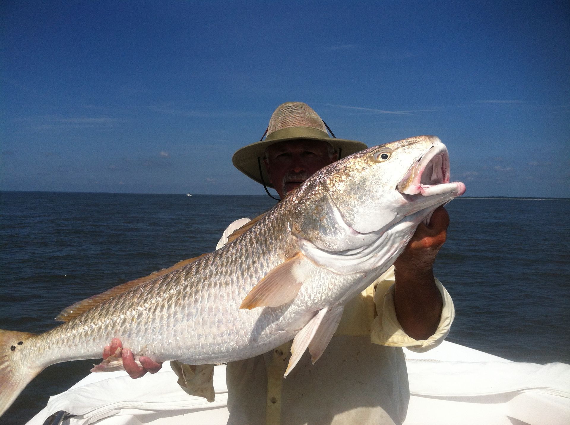 Man holding a large redfish with open mouth on a boat, blue water and sky background.