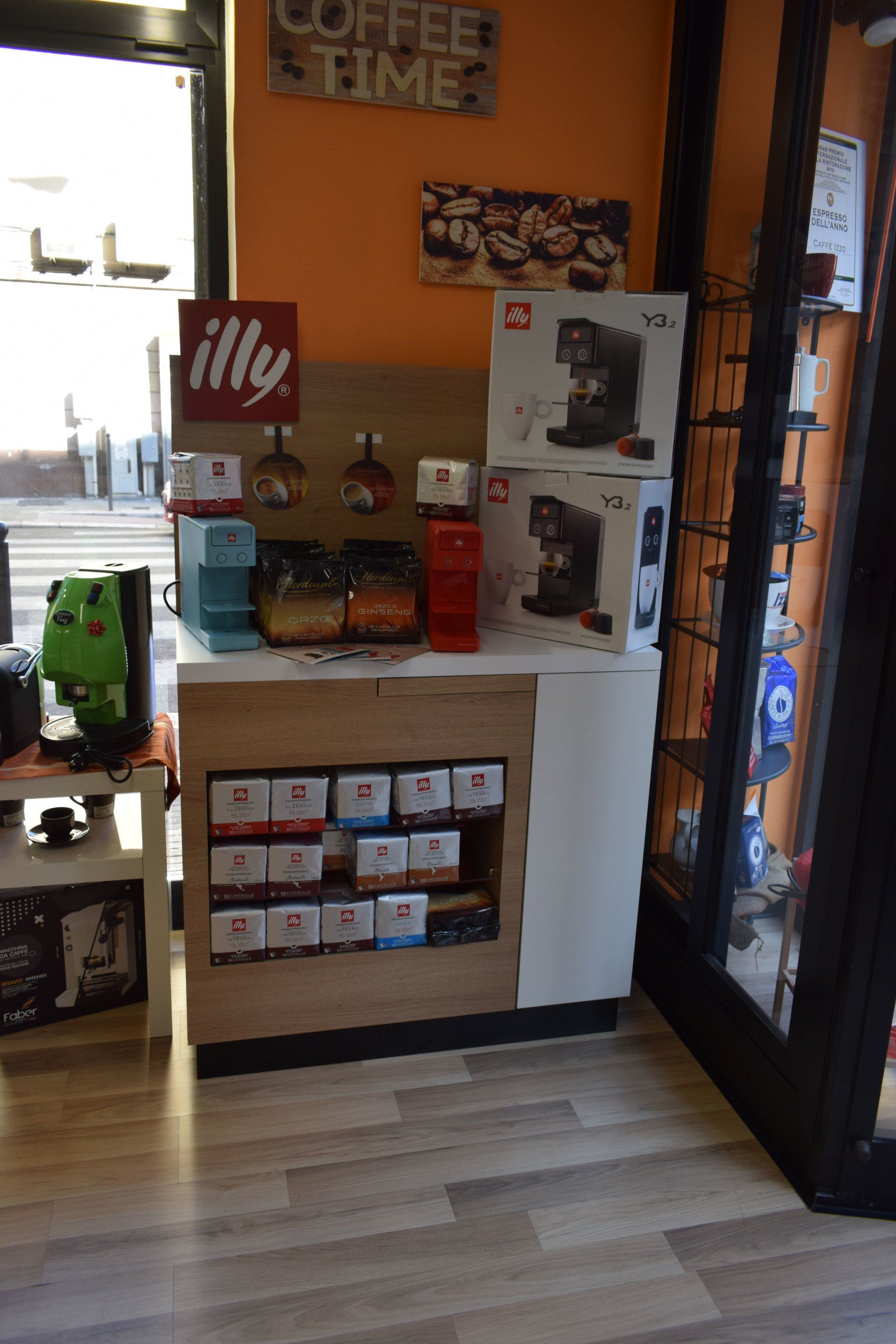 Esposizione di prodotti da caffè in un bar; insegna Illy, macchine da caffè e cialde di caffè sugli scaffali, bancone in legno, parete arancione.