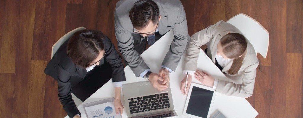 A top-down view of three people in business clothes at a table looking at a laptop.