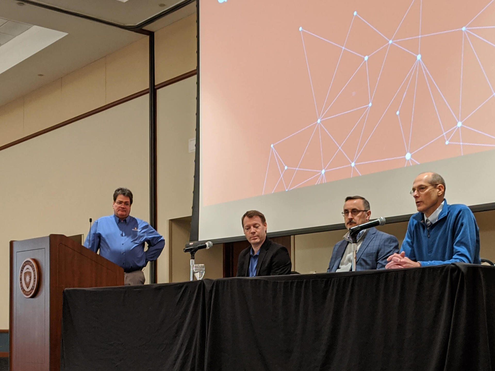 Three men seated at a table, one of them is speaking into a microphone. A man is at a podium and looking at him.