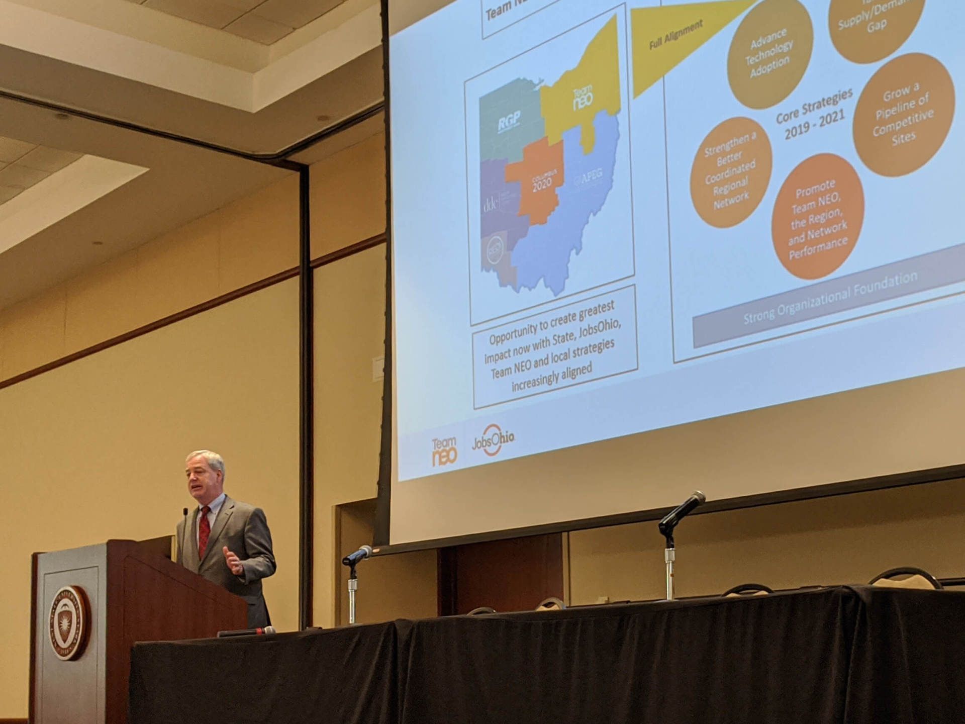 A man speaking into a microphone. A presentation slide is projected behind him.