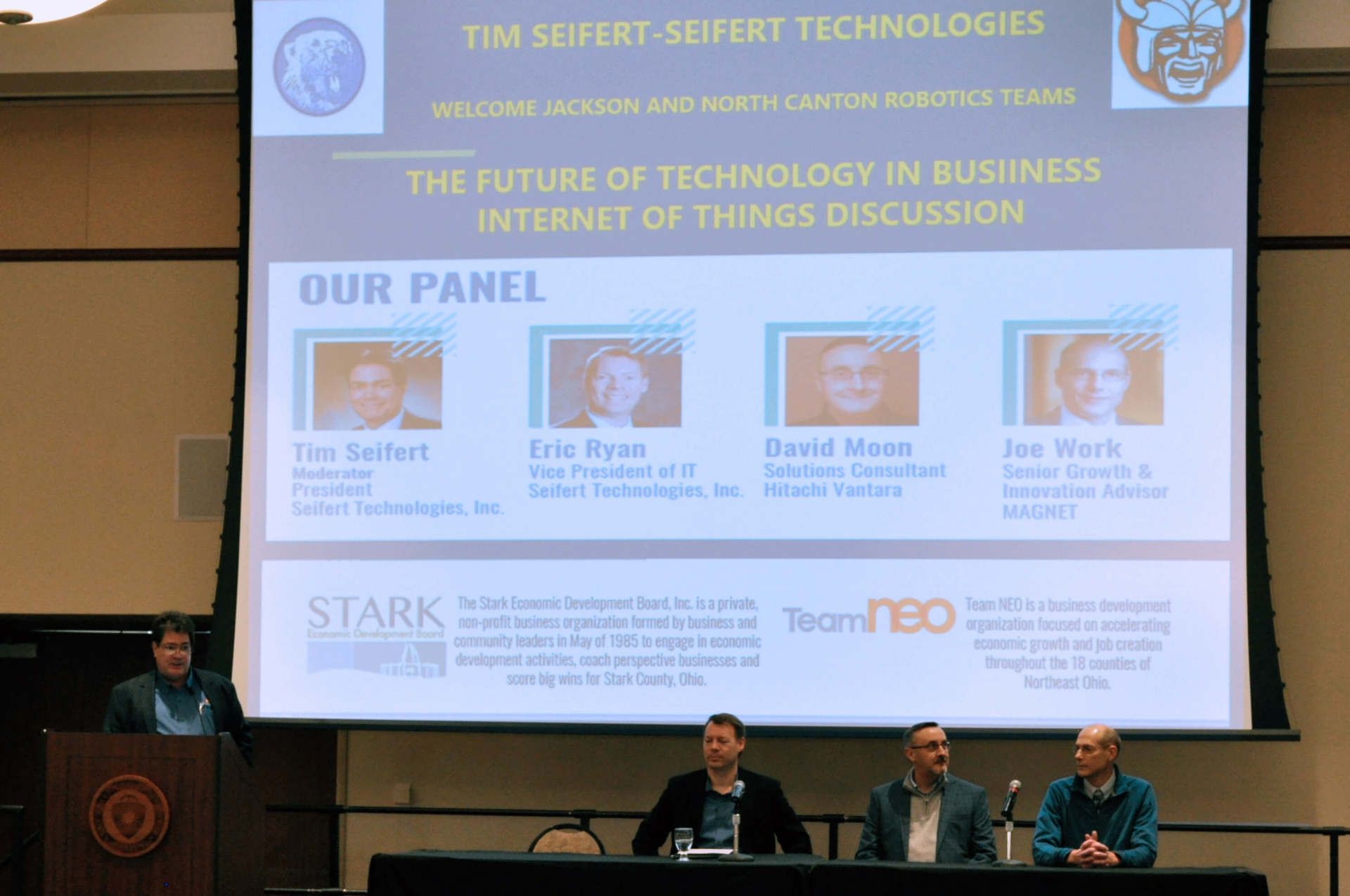 Three men seated at a table, another is speaking into a microphone. A presentation slide is projected behind them.