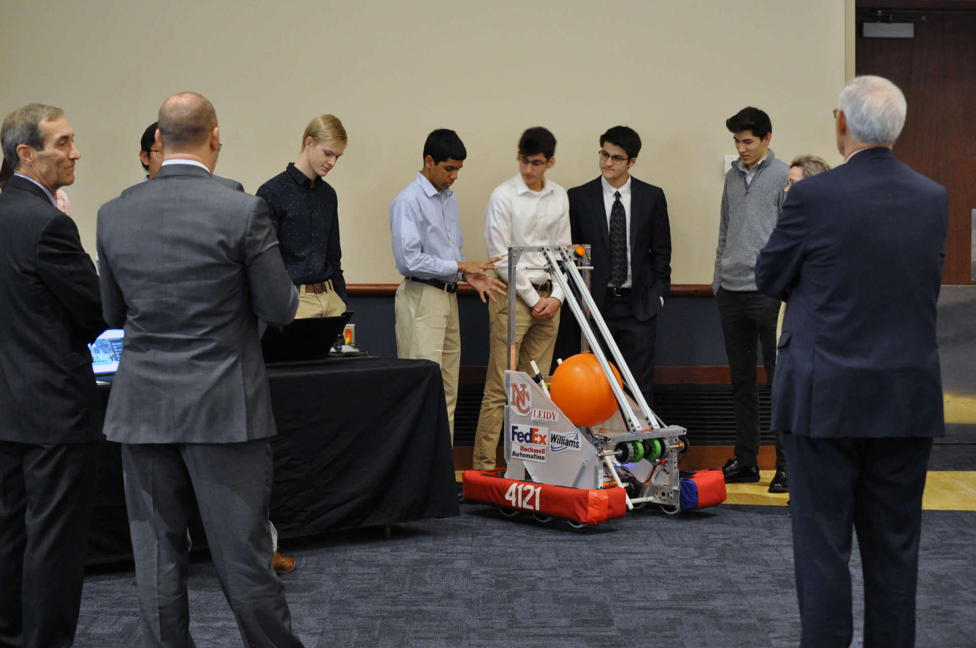 A couple of students are showcasing a robot that holds a large orange ball. A few other people are gathered around.