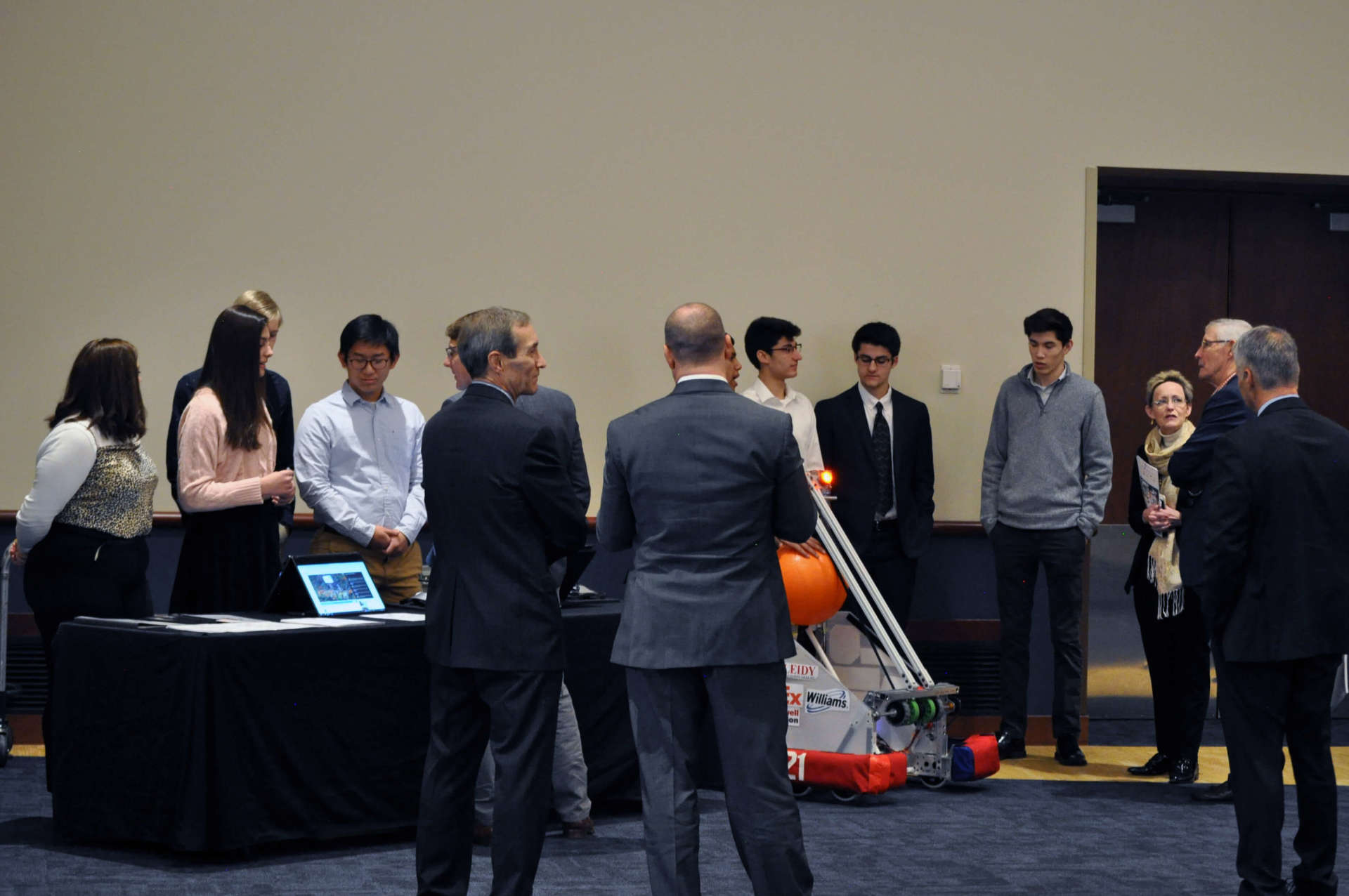 A couple of students are showcasing a robot that holds a large orange ball. A few other people are gathered around.