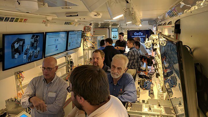 Nine people inside a trailer with various computers and equipment on display inside of it.