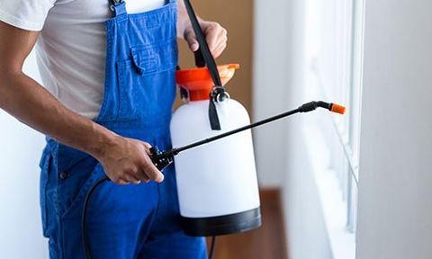 Un uomo con una tuta da lavoro con una tanica in plastica a tracolla e un spruzzino in mano 