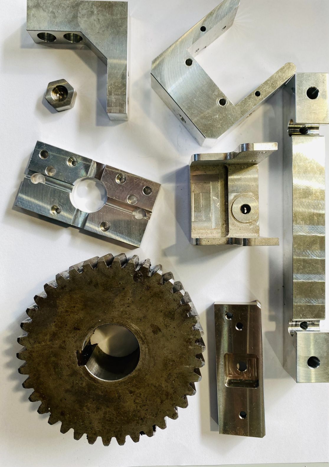 Parti metalliche di macchinari: ingranaggi, staffe e blocchi di varie forme e dimensioni, sparsi su una superficie bianca.