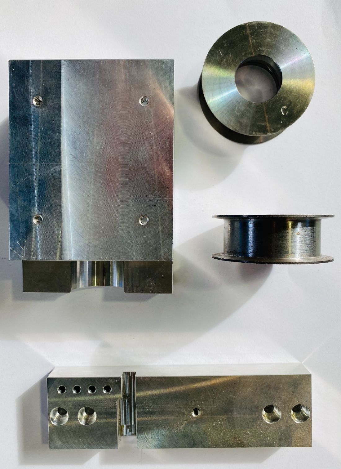 Diverse parti metalliche lavorate esposte su una superficie bianca, tra cui blocchi rettangolari e bobine.