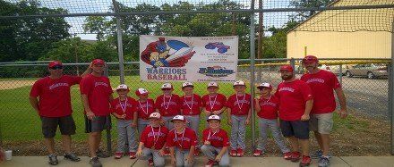 Group in red shirts and caps posing in front of a banner by a fenced sports field.