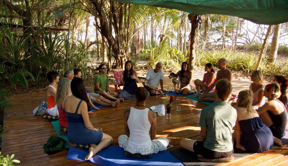 Group of people seated in a circle on mats, outdoors. Light shines through trees.