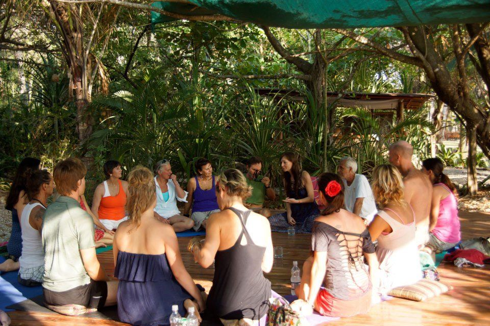 A group sits in a circle outdoors, interacting. Sunlight filters through trees. People wear casual clothing.