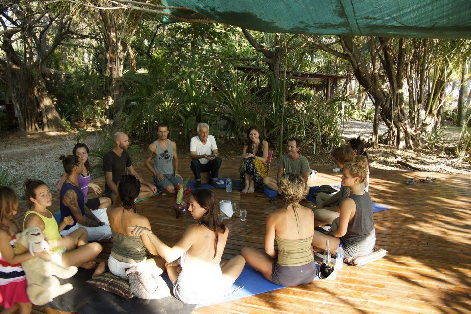 A group seated in a circle outdoors, beneath trees and a canopy. People are interacting.