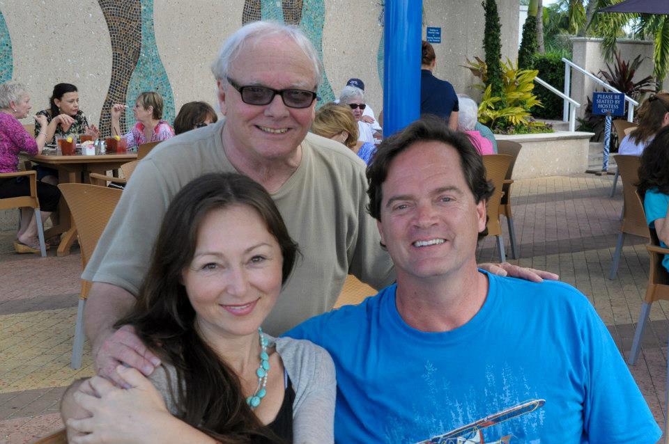 Three people smiling, posed outside near a light-colored wall, with others in the background.