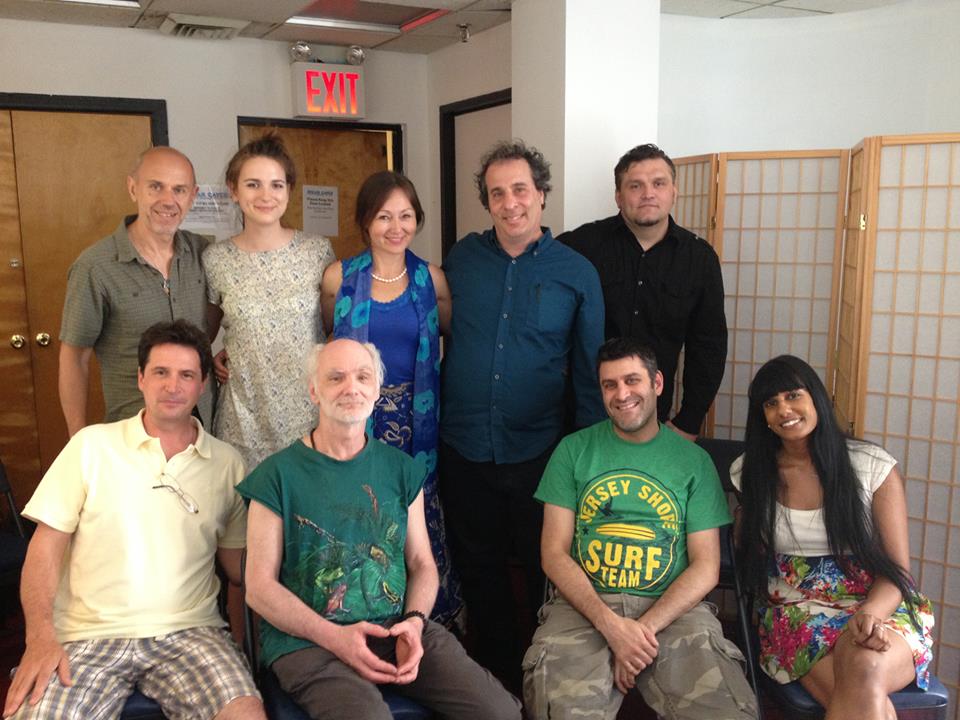 Group of people posing indoors; various skin tones, casual clothing. Some smiling, wood-paneled room.