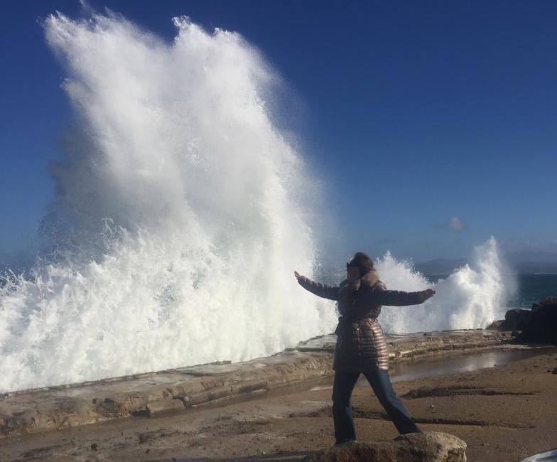 Person stands with arms outstretched, facing large wave crashing on rocky coast. Blue sky.