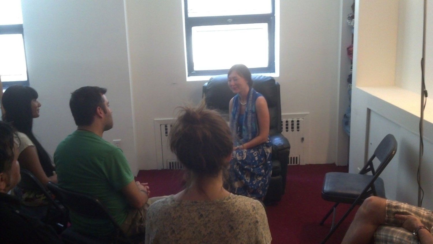 Woman in patterned dress sits, speaking to seated group indoors. Light from windows.