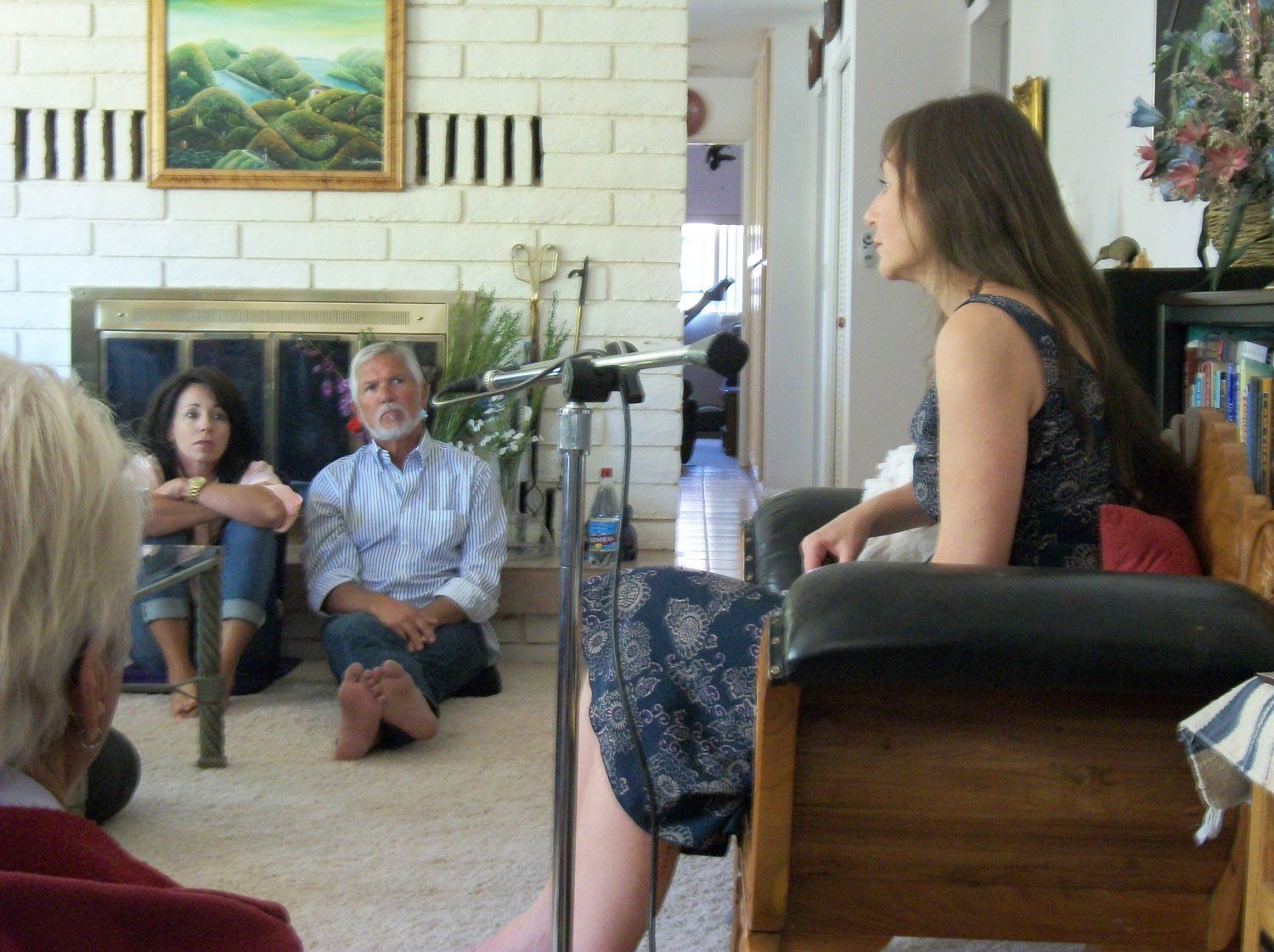 Woman in floral dress speaks to seated group in a living room.