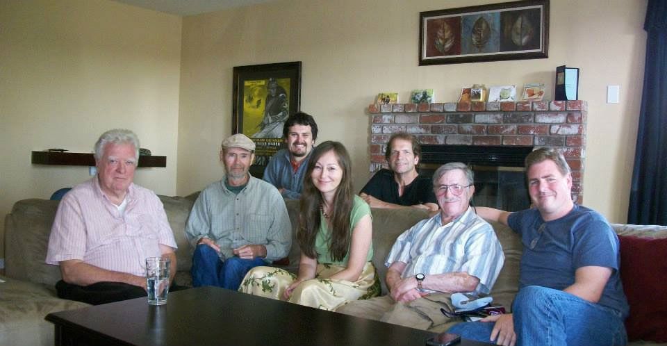 Group of seven people seated on a couch in a living room; smiling, looking at the camera.