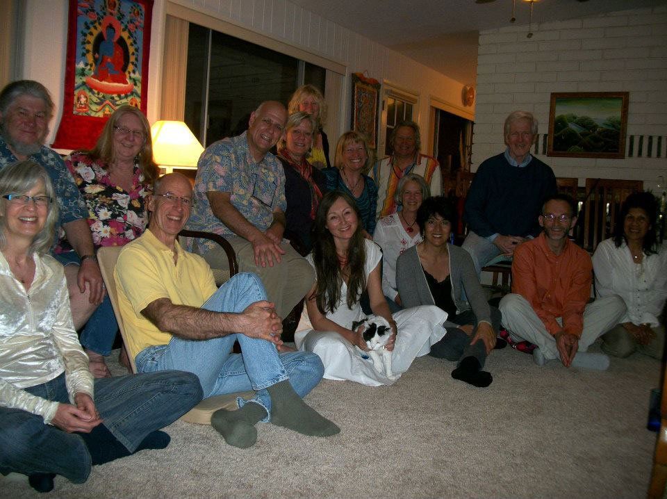 Group of people seated on a floor and chairs indoors. Some smiling. Buddha tapestry and artwork visible.