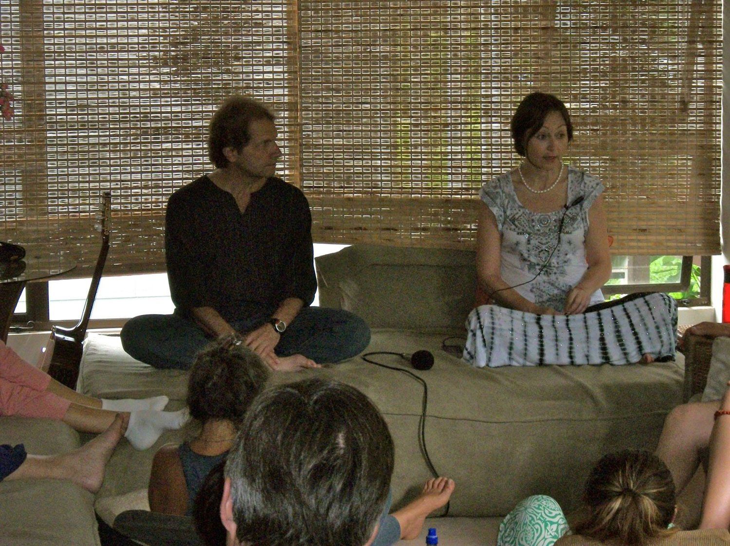 Two people seated cross-legged on a cushion, speaking to a seated audience, inside a bamboo-walled structure.