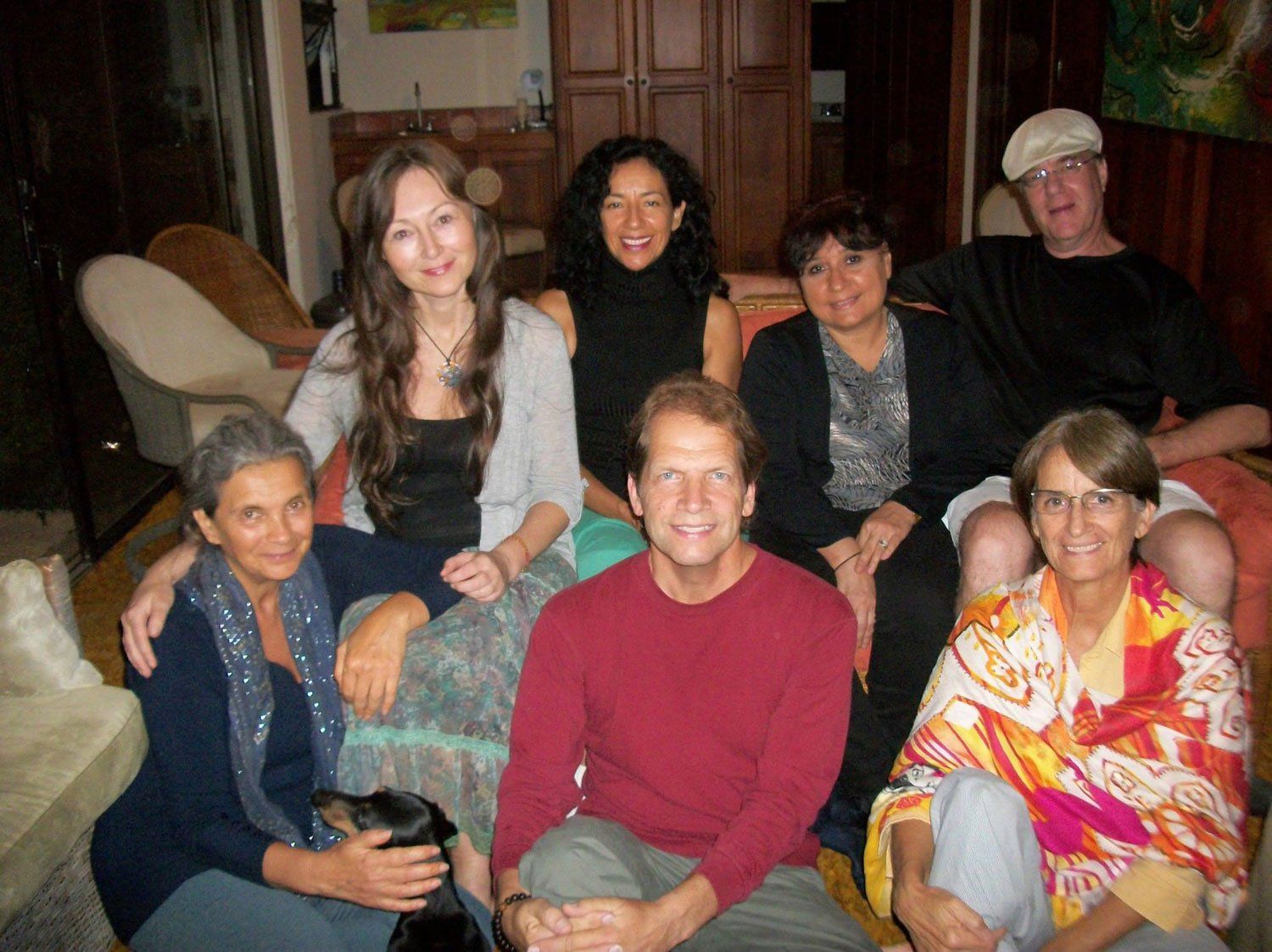 Group of people smiling, indoors. Women, men, and a small dog are together on a couch and floor.