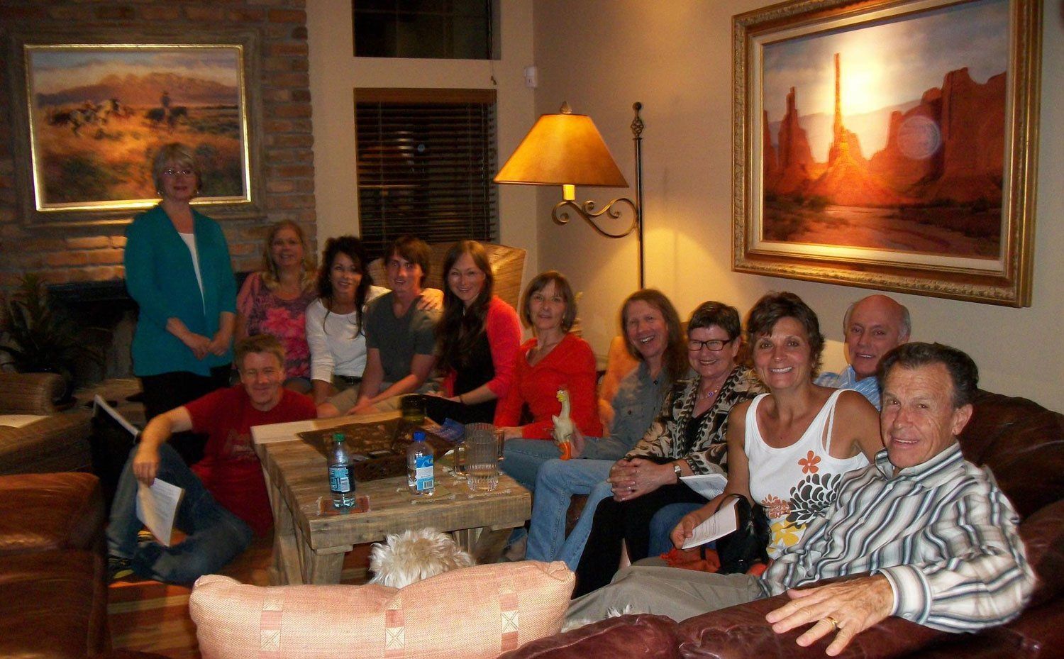 Group of people gathered in a living room, some seated on a couch, others standing, near a coffee table.