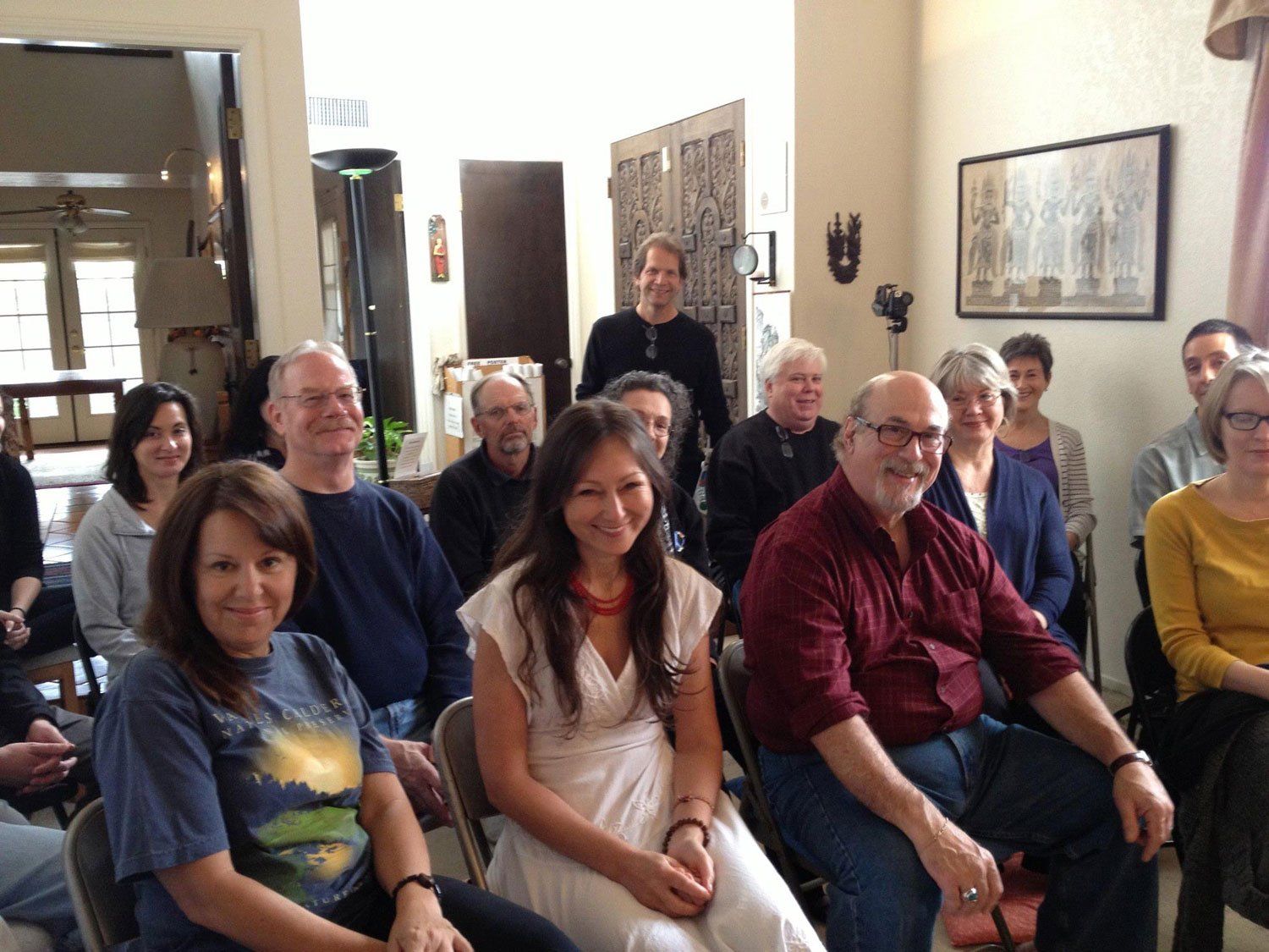 A group of people sitting in chairs indoors, smiling and looking forward. Some appear to be listening.