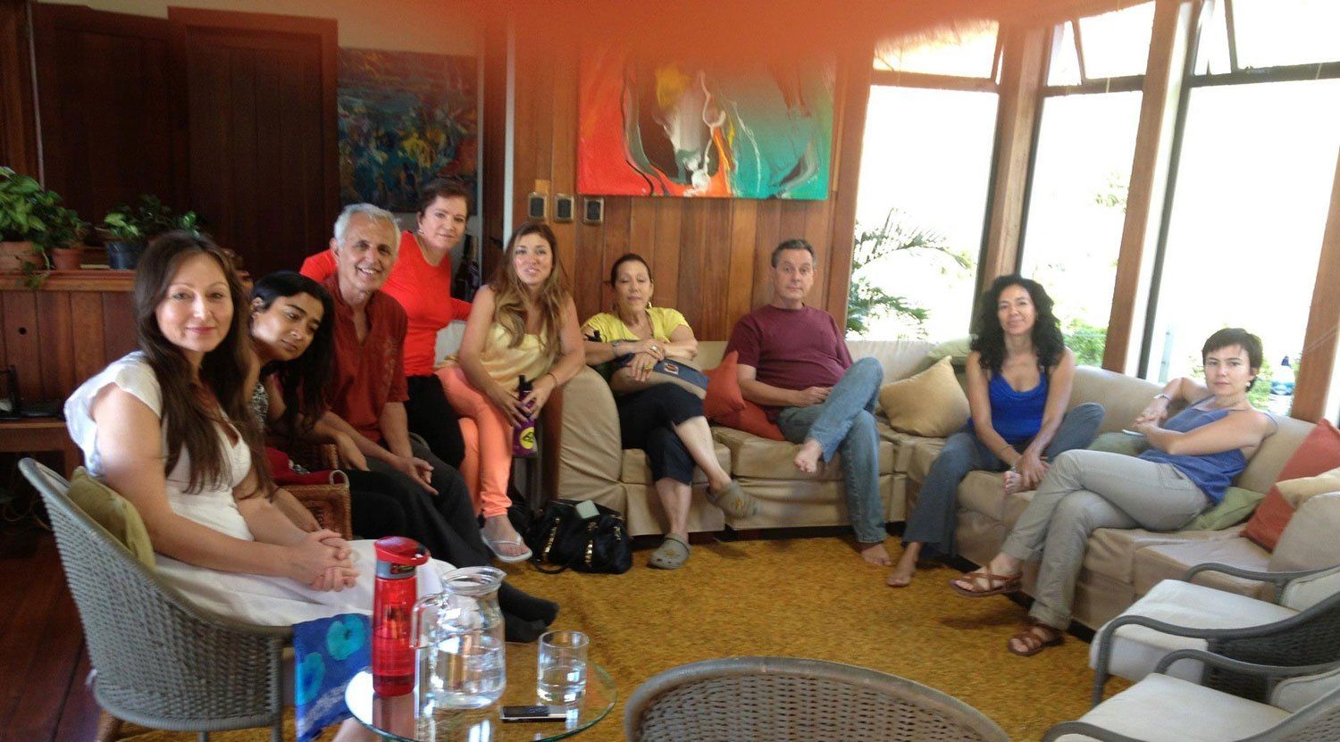 A group of people sitting in a living room, some smiling. There's a couch, art, and plants.
