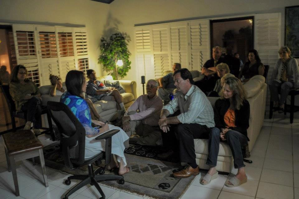 People gathered indoors, seated or lounging, listening to two individuals speaking. Dim lighting, shuttered windows.